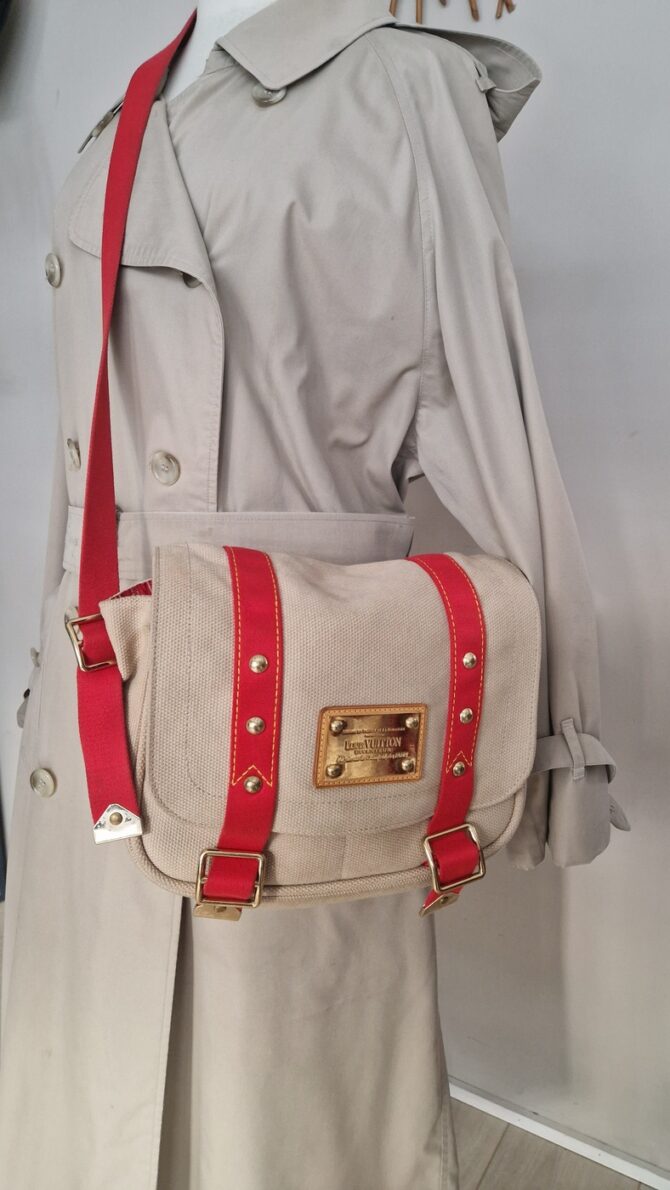 Sac Louis Vuitton Antigua Besace PM en toile beige avec sangles rouges et plaque Inventeur dorée Il est presenté porté EN : Louis Vuitton Antigua Besace PM in beige canvas with red straps and gold Inventeur plaque